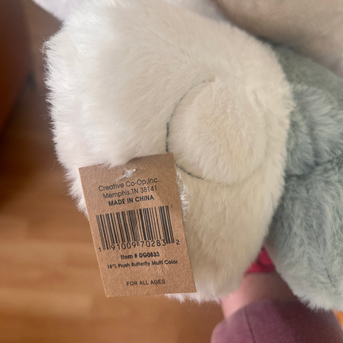 Close-up of a plush stuffed animal showing soft white and pale green fur with a brown product tag attached. The tag reads Creative Co-Op Inc.
