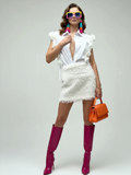 Woman in a white outfit with ruffled top and fringed skirt, wearing pink high boots and colorful sunglasses, holding an orange handbag.