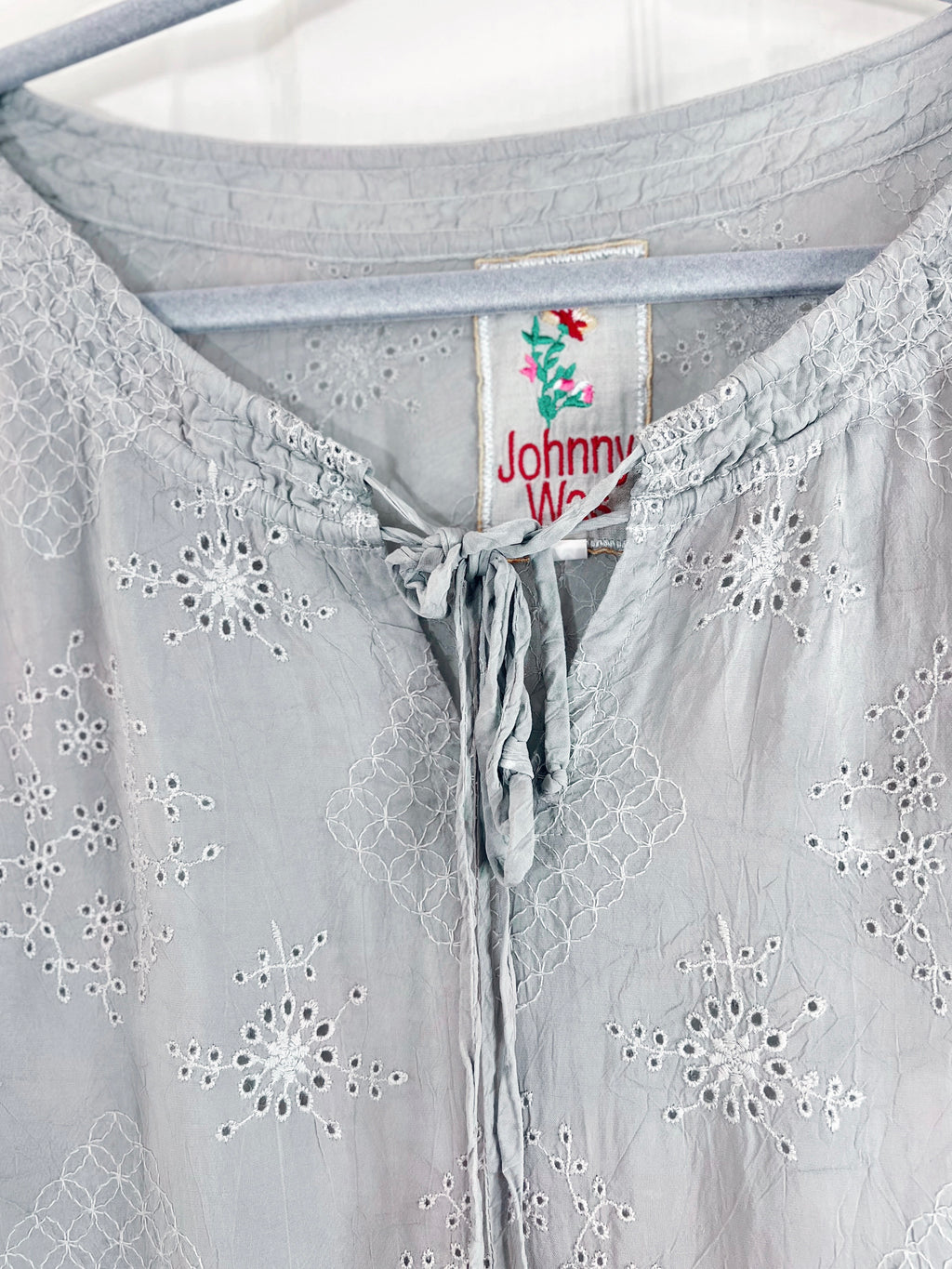 Close-up of a light gray eyelet blouse hanging on a hanger, showing embroidered floral patterns and a drawstring neckline. The inner neck label reads Johnny Walker.