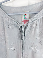 Close-up of a light gray eyelet blouse hanging on a hanger, showing embroidered floral patterns and a drawstring neckline. The inner neck label reads Johnny Walker.