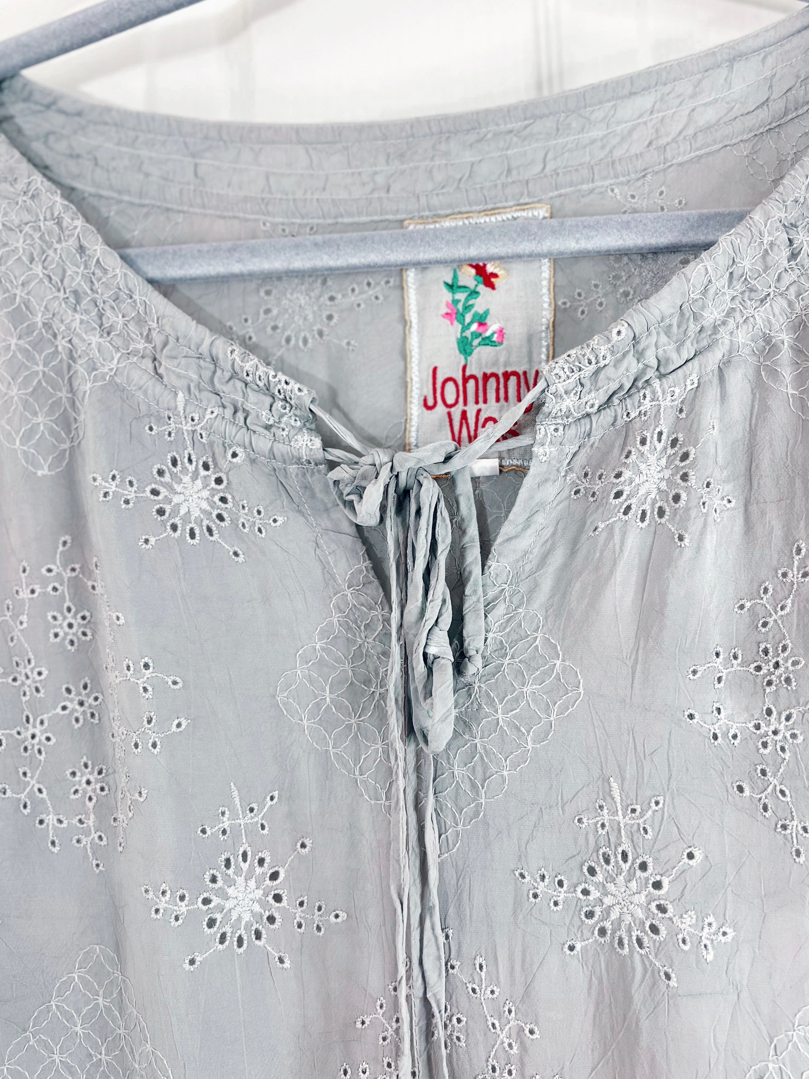 Close-up of a light gray eyelet blouse hanging on a hanger, showing embroidered floral patterns and a drawstring neckline. The inner neck label reads Johnny Walker.