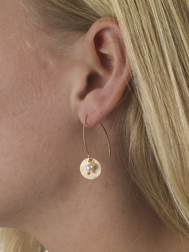 Gold hoop earring with a pearl drop worn by a person with blonde hair.