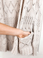 Close-up of a person's hand reaching into a light beige open-front knit cardigan with textured lace patterns and a pocket.