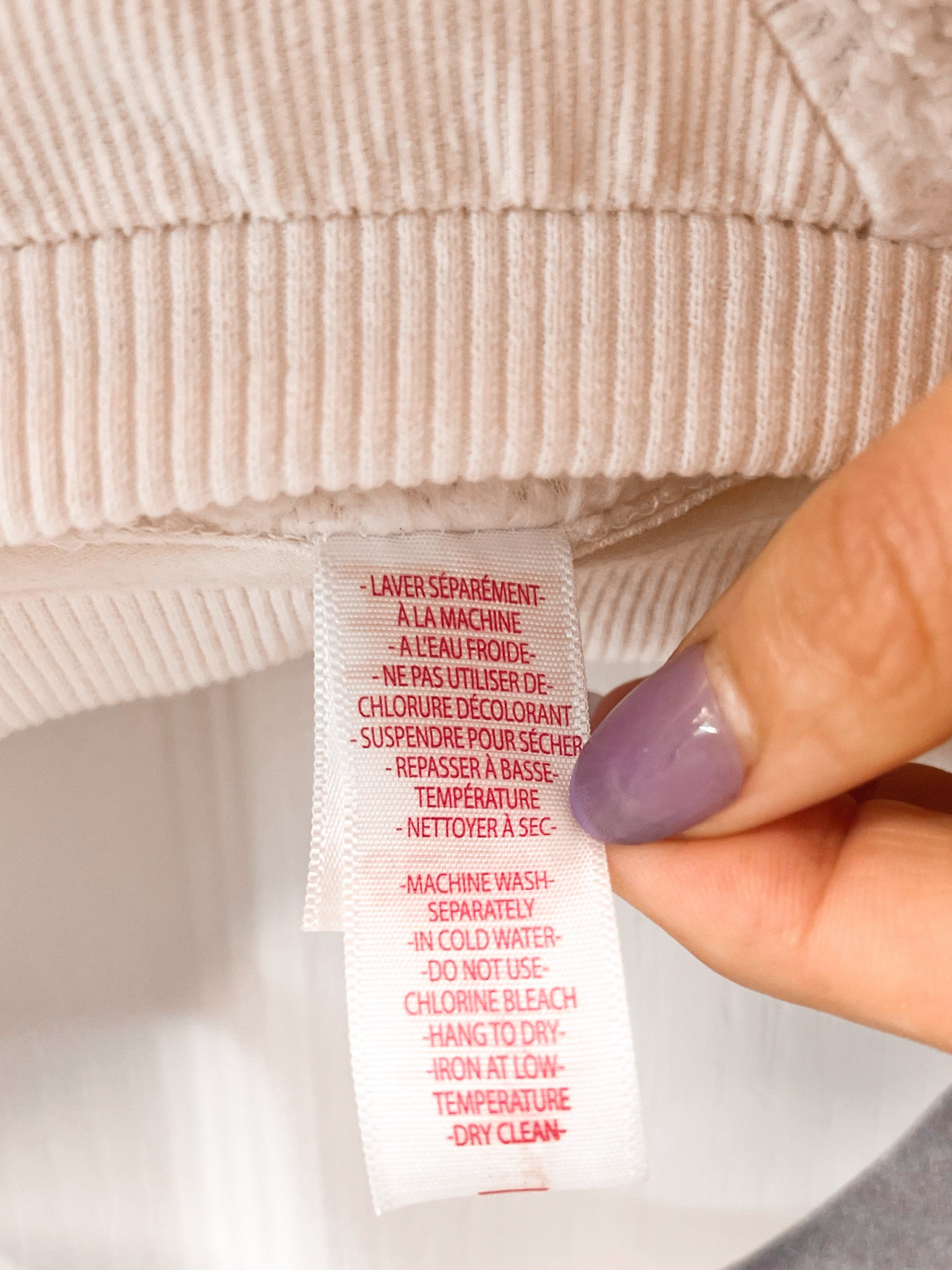 A close-up of a person's hand holding a white care label with red text attached to the ribbed collar of a light beige or cream knit garment, with a gray hanger visible at the top.