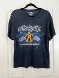 A black washed short-sleeve t-shirt hanging on a black hanger against a white paneled door. The shirt features a large graphic print for the band 'Aerossmith' with the text 'Devices Are Wild' and a central gold 'A' logo surrounded by blue wings.
