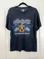 A black washed short-sleeve t-shirt hanging on a black hanger against a white paneled door. The shirt features a large graphic print for the band 'Aerossmith' with the text 'Devices Are Wild' and a central gold 'A' logo surrounded by blue wings.