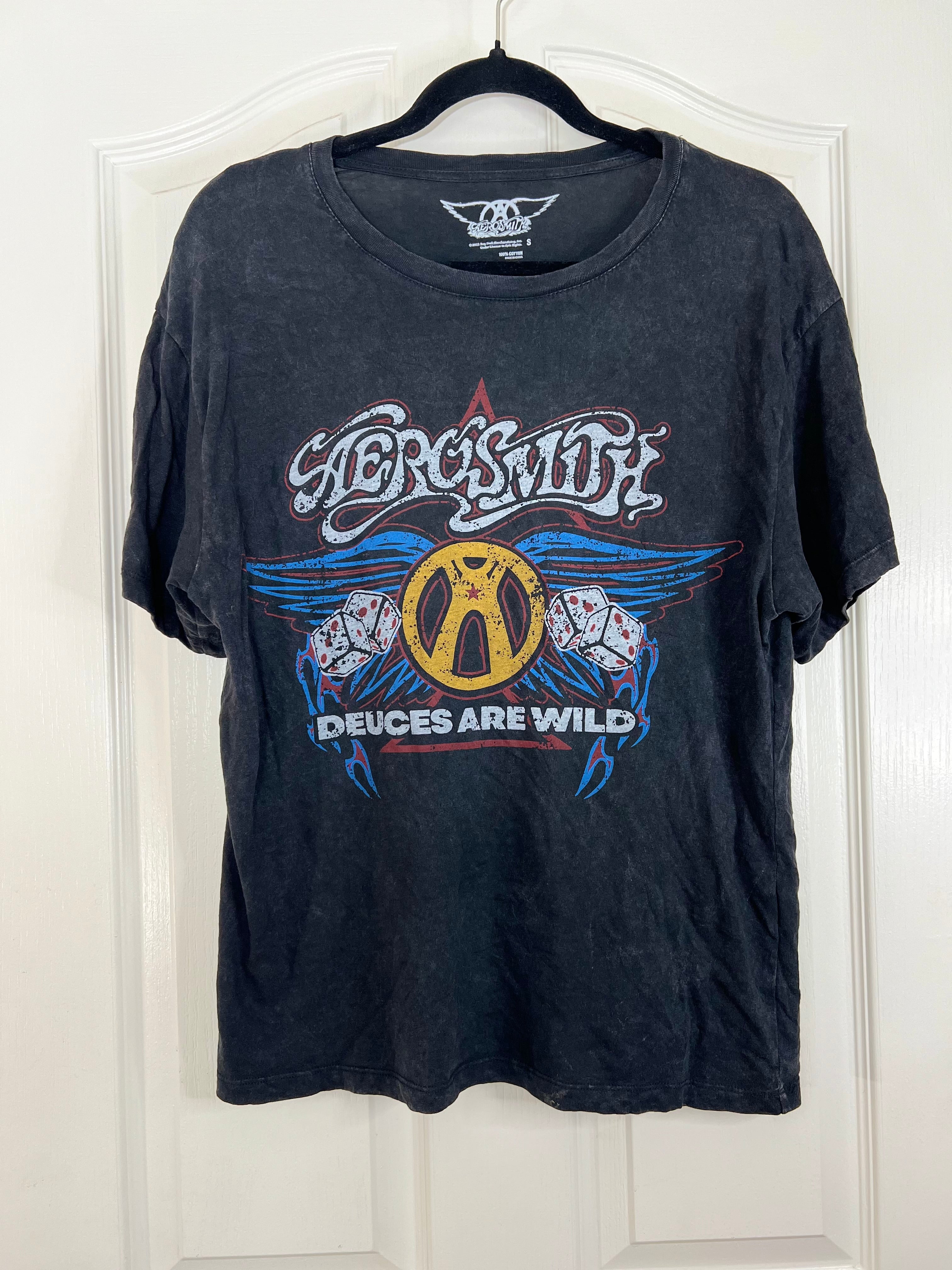 A black washed short-sleeve t-shirt hanging on a black hanger against a white paneled door. The shirt features a large graphic print for the band 'Aerossmith' with the text 'Devices Are Wild' and a central gold 'A' logo surrounded by blue wings.