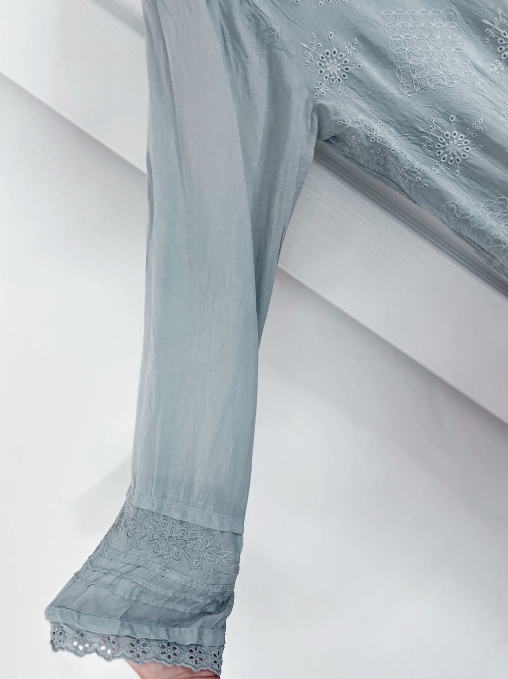 A close-up of a light blue-gray sheer fabric garment with embroidered details and a lace-trimmed hem, draped over a white bench.