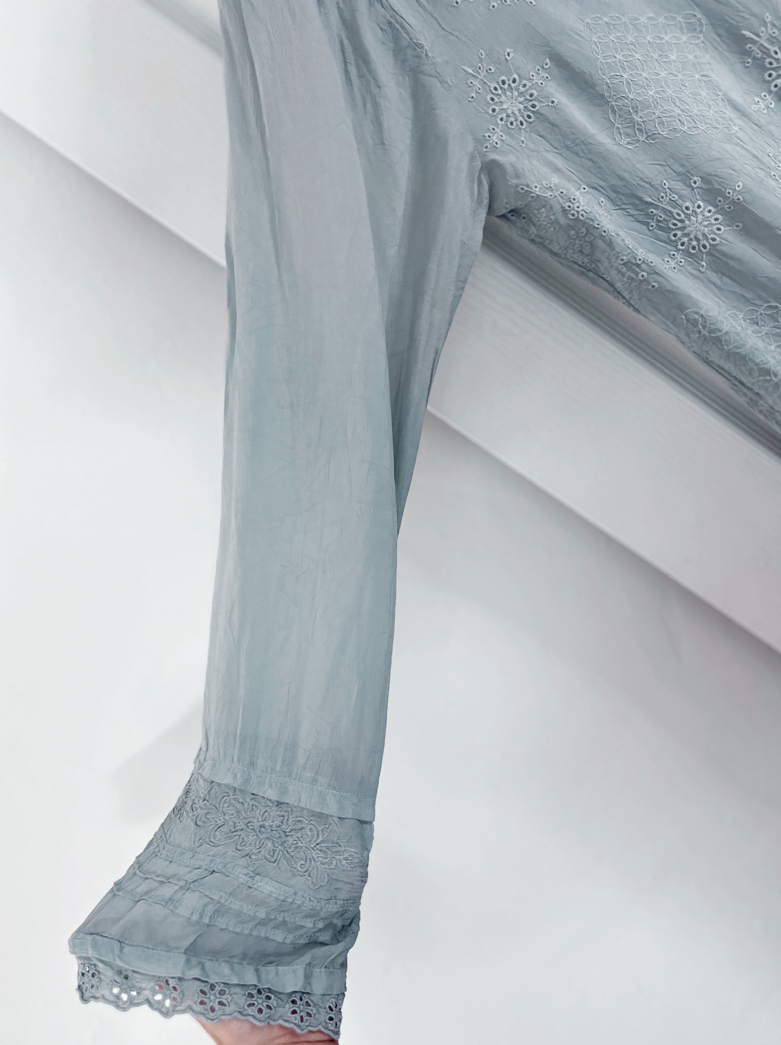 A close-up of a light blue-gray sheer fabric garment with embroidered details and a lace-trimmed hem, draped over a white bench.