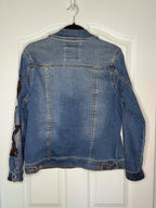 A blue denim jacket hanging on a black hanger against a white paneled door, showing the back view with button details and contrast stitching.