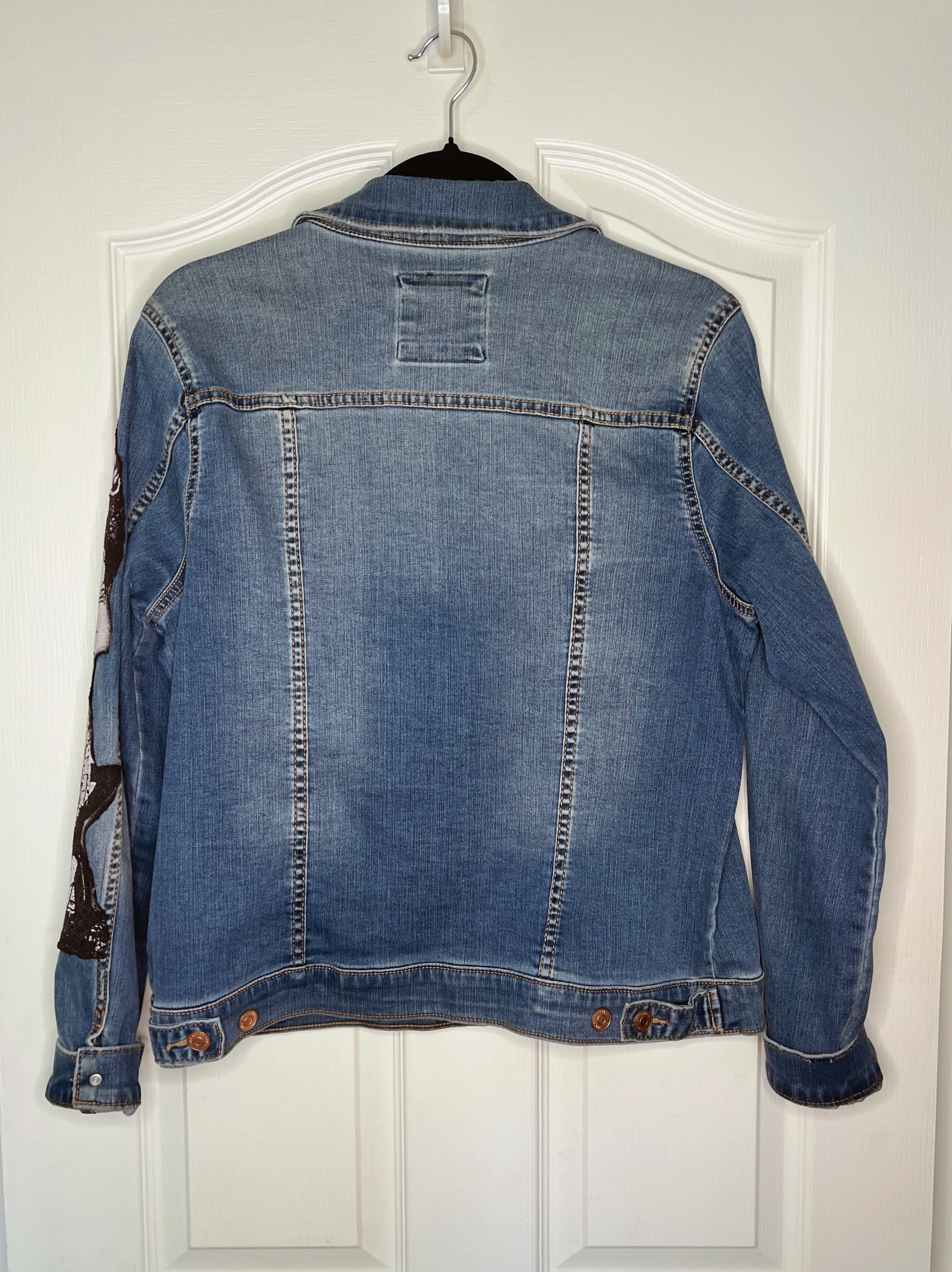 A blue denim jacket hanging on a black hanger against a white paneled door, showing the back view with button details and contrast stitching.