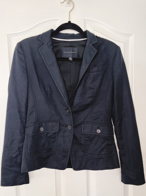 A navy blue women's blazer jacket hanging on a black hanger against a white paneled door. The jacket features notch lapels, a two-button front closure, and two front flap pockets with silver-tone buttons.