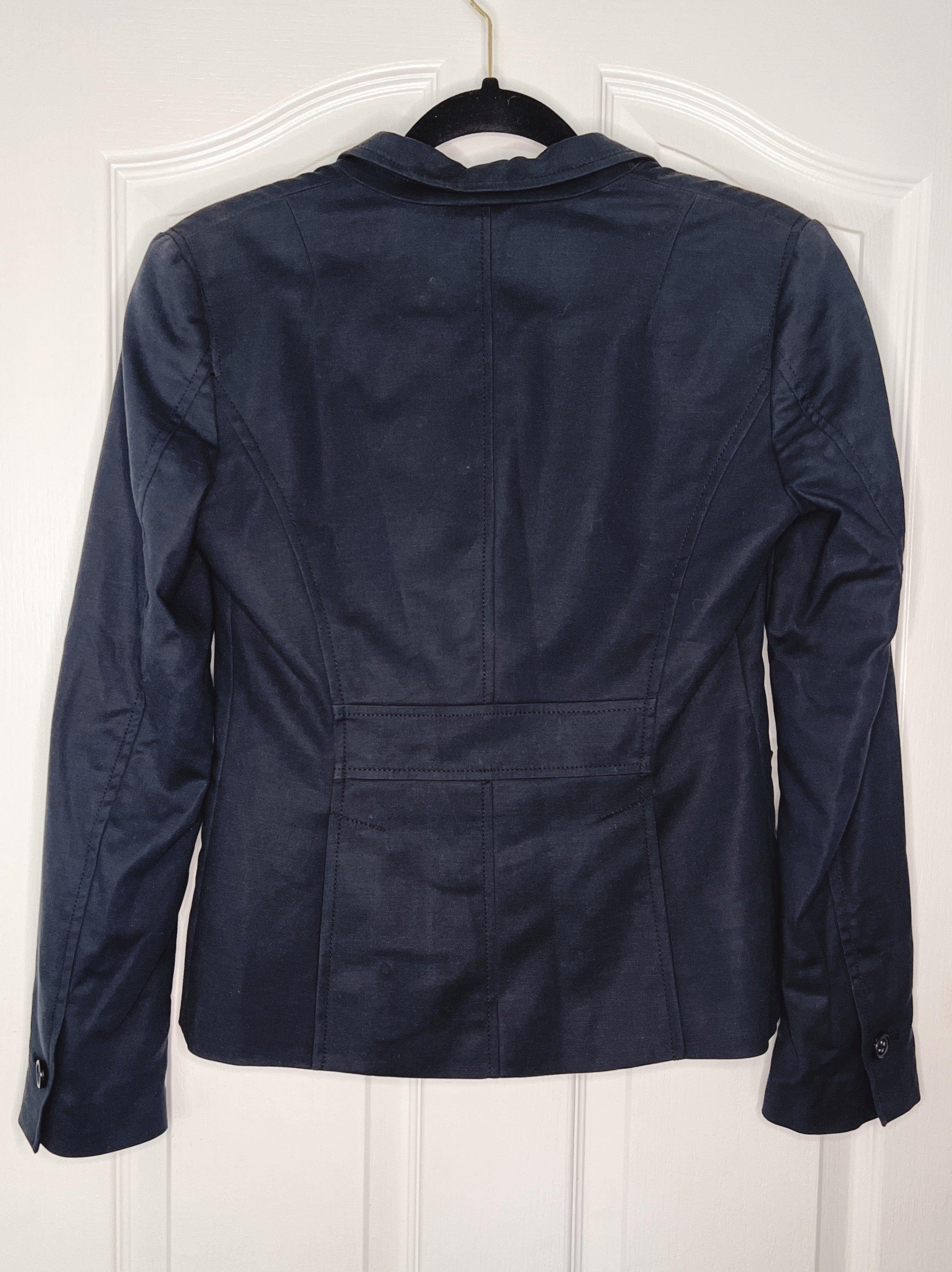 A dark navy blue cropped jacket with a notched lapel collar, shown from the back hanging on a black hanger against a white paneled door.