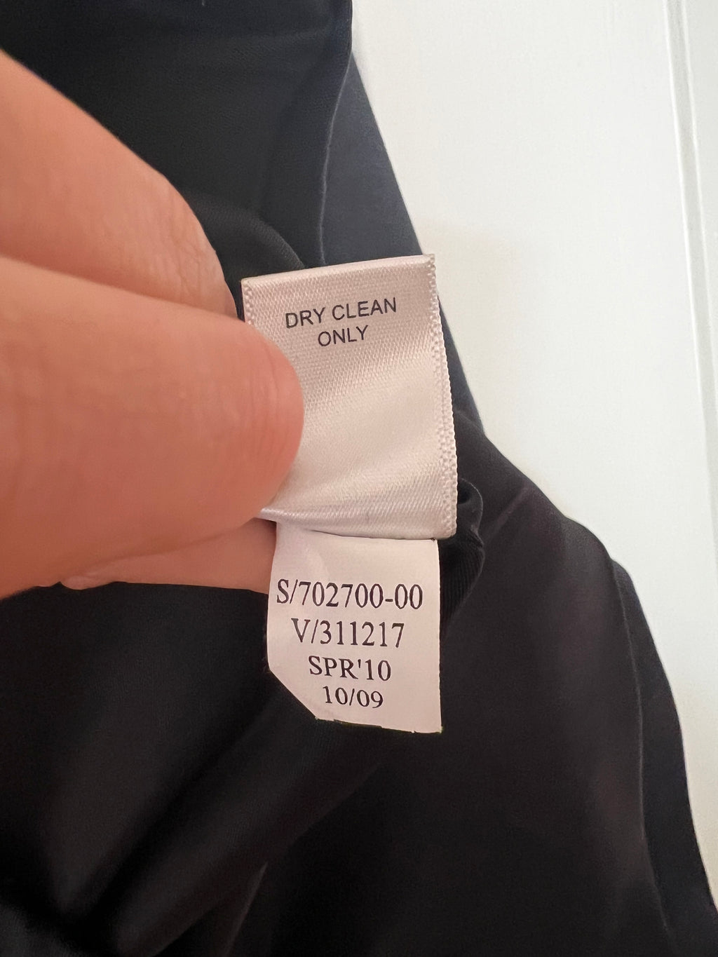 Close-up of a black garment's interior neck label being held by a finger, showing a white satin tag with black printed text including 'DRY CLEAN ONLY' and a style code.