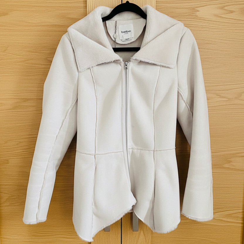 A beige, waist-length Bebe coat with an uneven hem and raw seam detailing, displayed on a hanger.