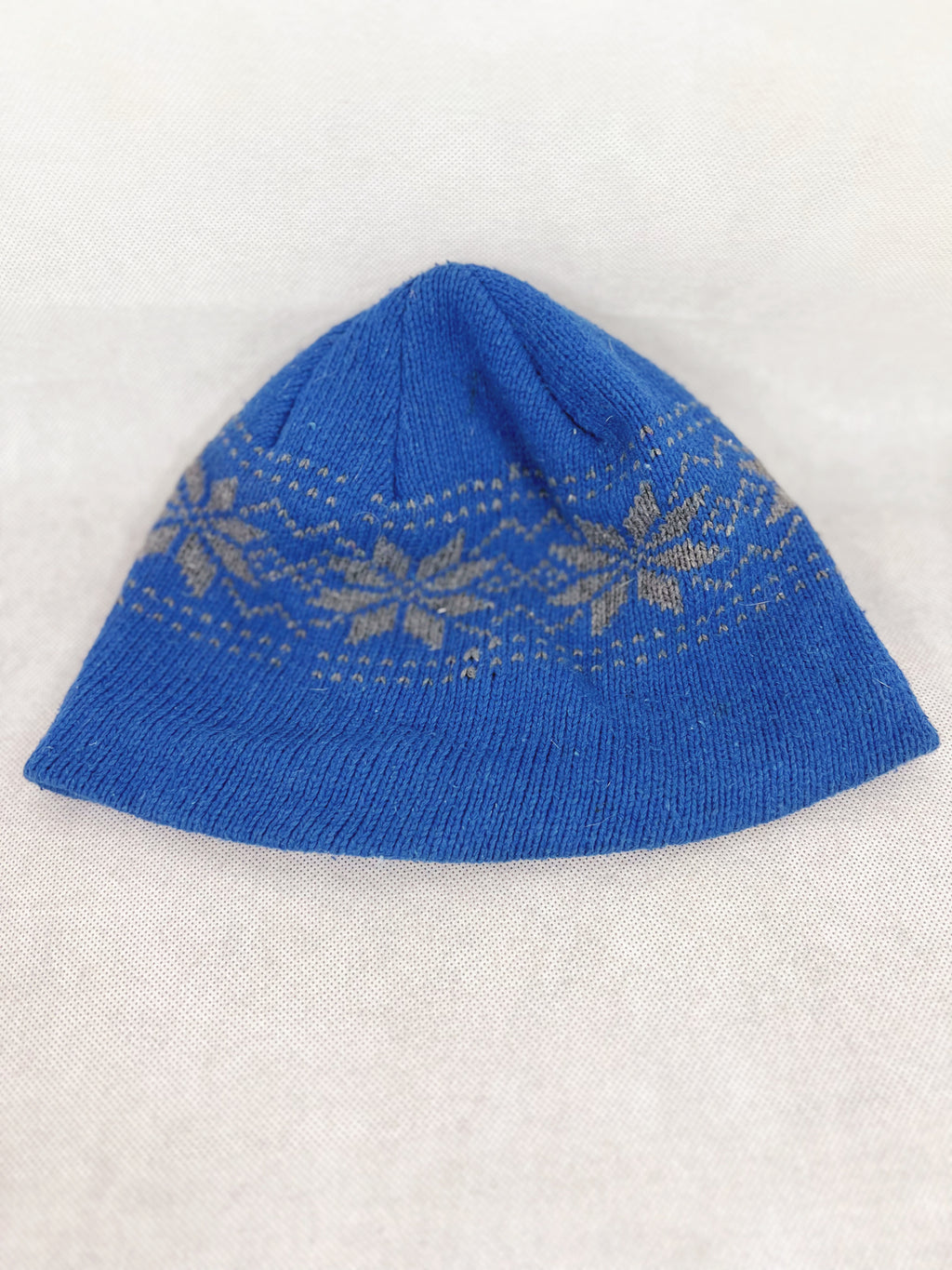 Back side of ablue knit beanie hat featuring a light beige Fair Isle snowflake pattern around the middle, displayed on a light beige textured fabric background.