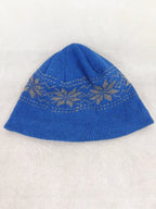 Back side of ablue knit beanie hat featuring a light beige Fair Isle snowflake pattern around the middle, displayed on a light beige textured fabric background.