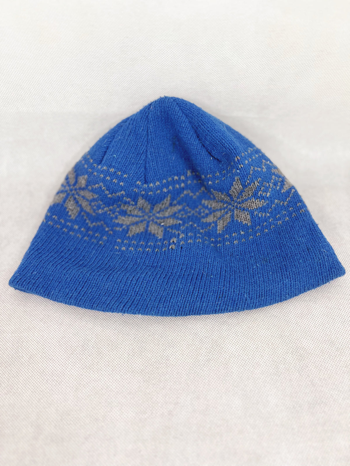 Back side of ablue knit beanie hat featuring a light beige Fair Isle snowflake pattern around the middle, displayed on a light beige textured fabric background.