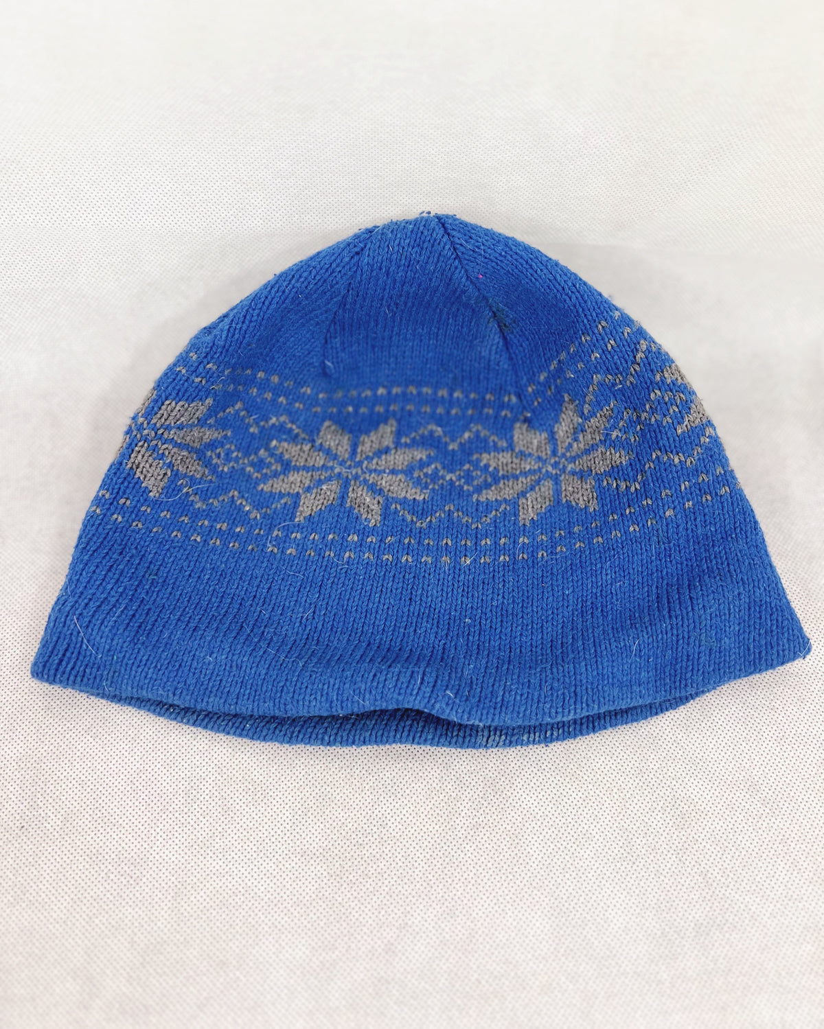 A front side blue knit beanie hat featuring a light beige Fair Isle pattern band across the upper half, laid flat on a light beige textured fabric background.