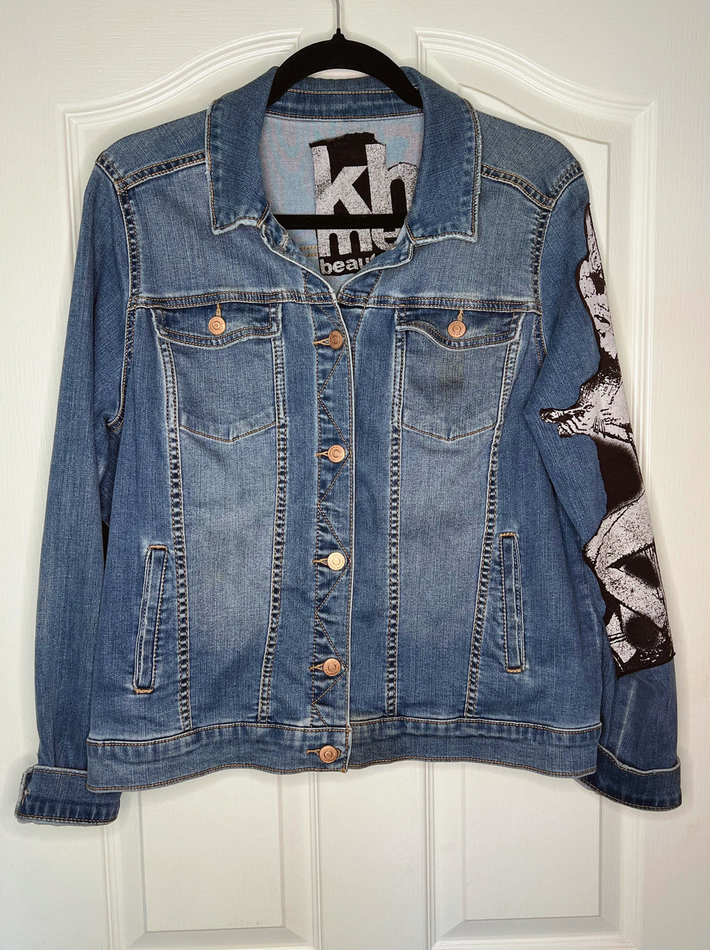 A blue denim jacket with a black and white graphic print on the right sleeve, hanging on a black hanger against a white paneled door.