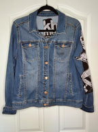 A blue denim jacket with a black and white graphic print on the right sleeve, hanging on a black hanger against a white paneled door.