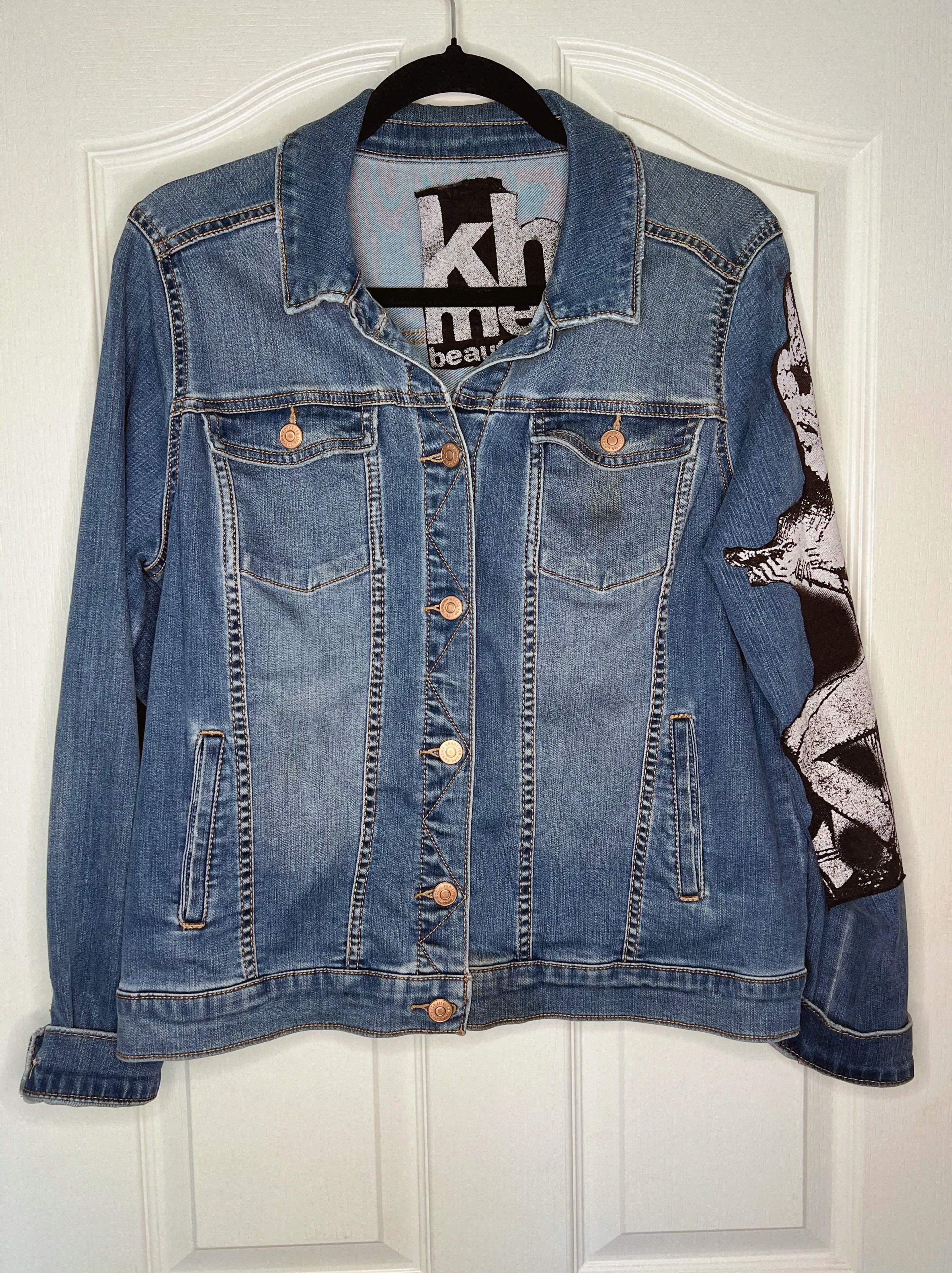 A blue denim jacket with a black and white graphic print on the right sleeve, hanging on a black hanger against a white paneled door.