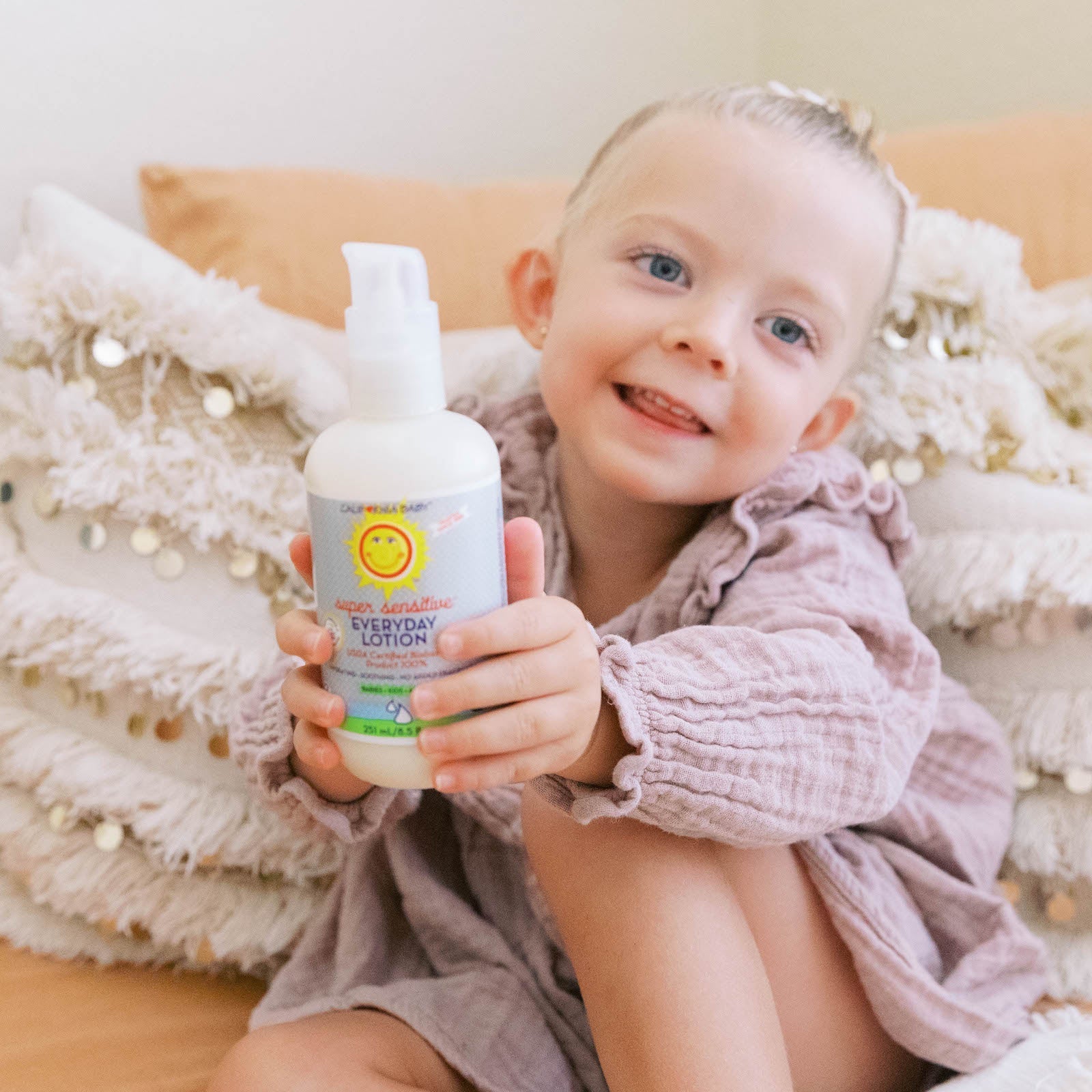 Child holding a bottle of lotion with a visible brand logo, sitting on a couch.