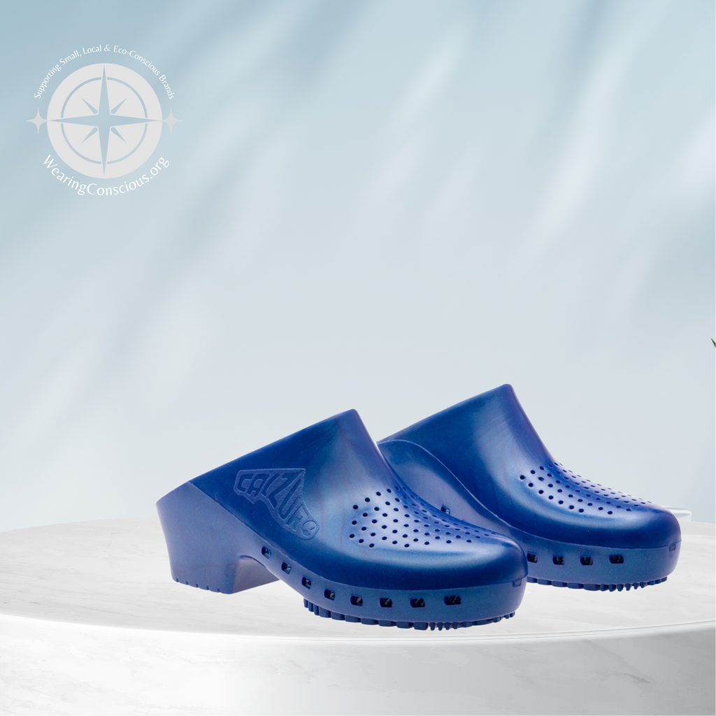 A pair of bright blue clog-style shoes with perforated details and low block heels, displayed on a white cylindrical pedestal against a light blue background with soft leaf shadows and a small green plant.