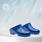 A pair of bright blue clog-style shoes with perforated details and low block heels, displayed on a white cylindrical pedestal against a light blue background with soft leaf shadows and a small green plant.