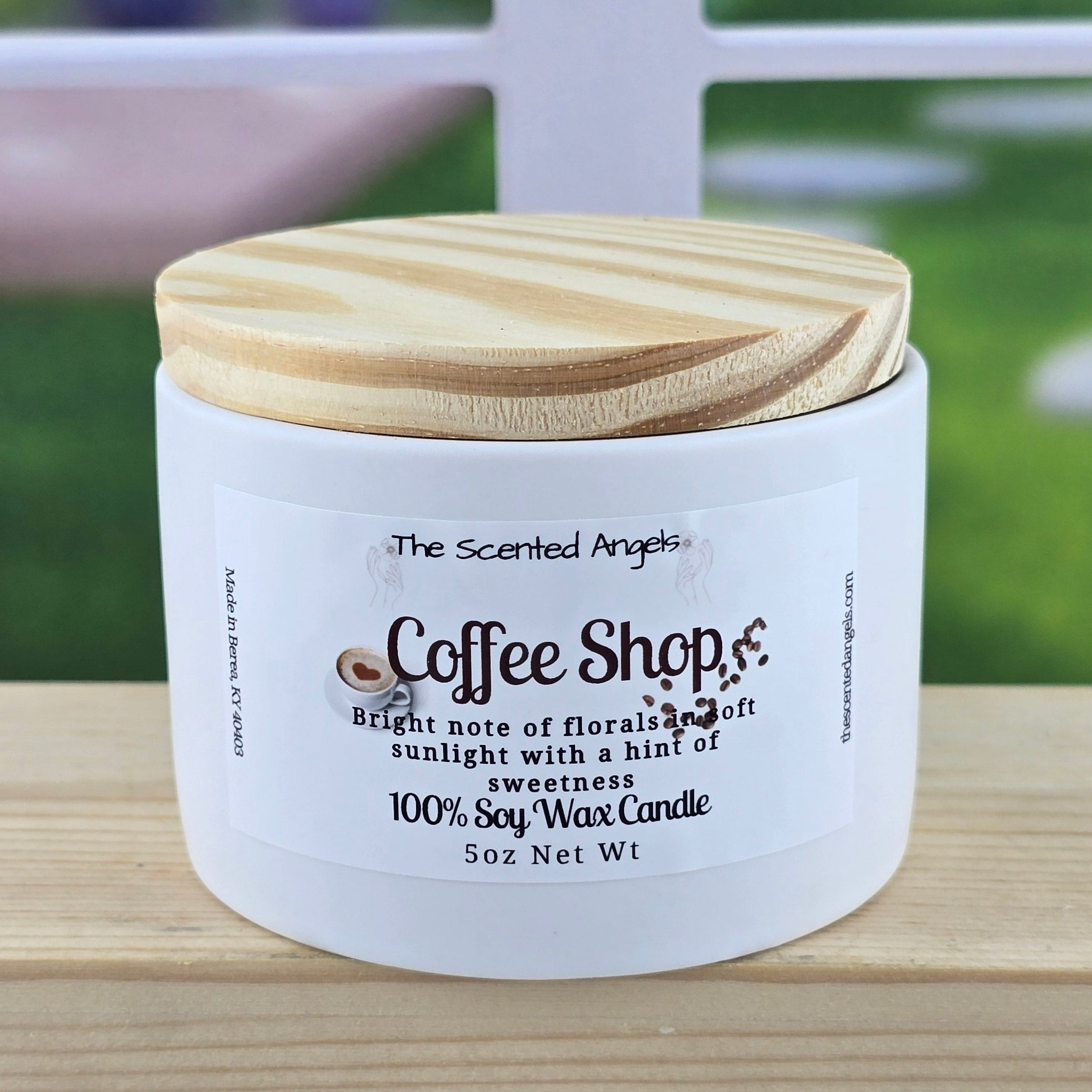 Candle jar labeled 'Coffee Shop' by 'The Scented Angel' on a wooden surface with a blurred outdoor background.