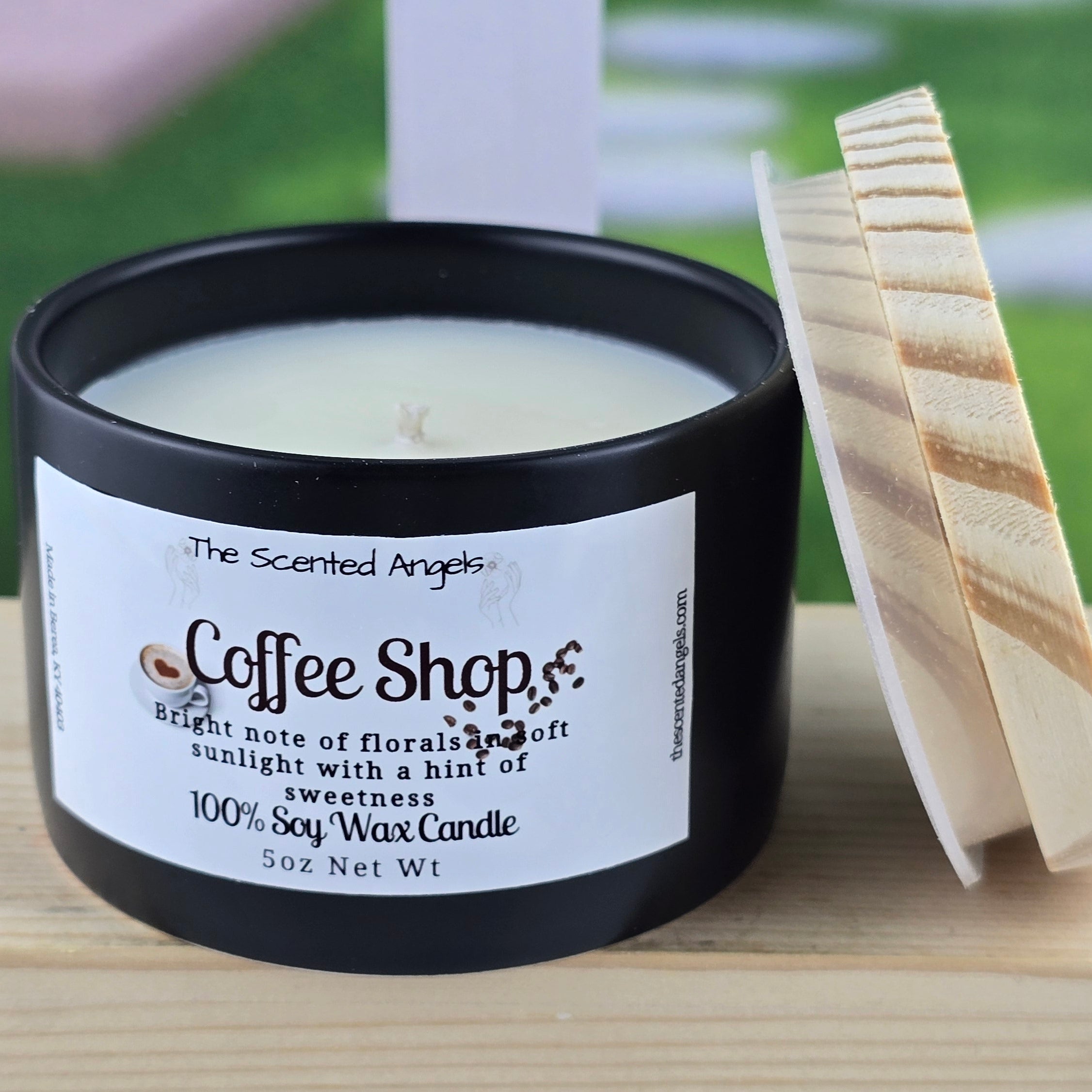 Candle labeled 'Coffee Shops' by 'The Scented Angel' on a wooden surface with a blurred outdoor background.