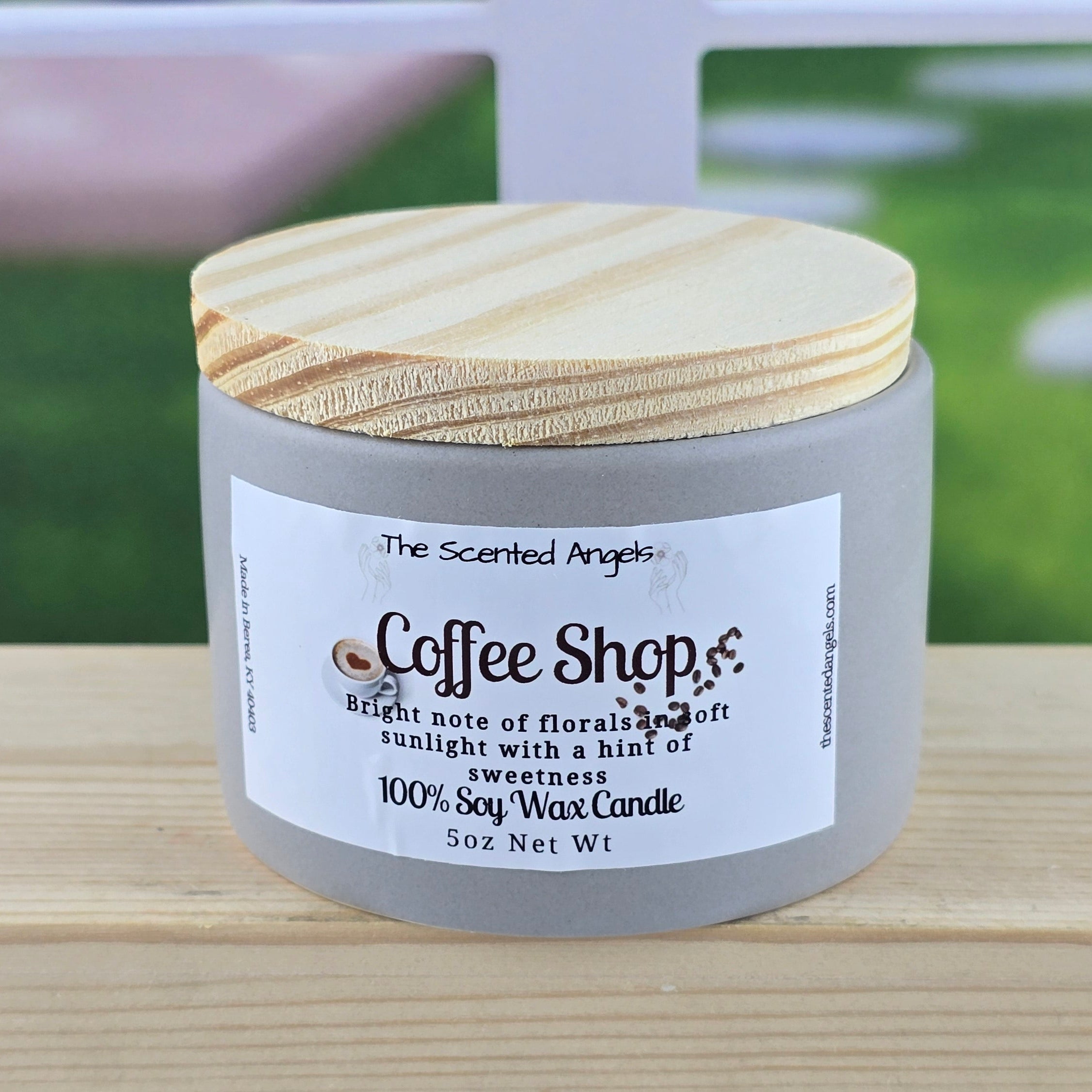 Candle labeled 'Coffee Shop' by 'The Scented Angel' on a wooden surface with a blurred outdoor background.