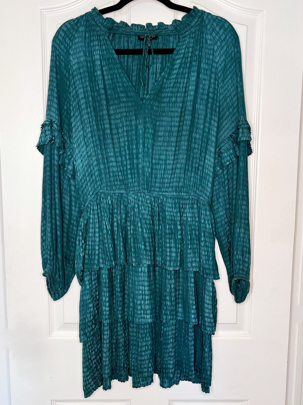 A teal green tiered long-sleeve dress with a textured, tie-dye-like pattern, hanging on a black hanger against a white paneled door.