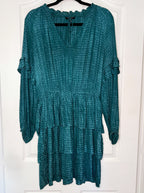 A teal green tiered long-sleeve dress with a textured, tie-dye-like pattern, hanging on a black hanger against a white paneled door.