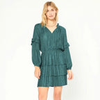 A woman modeling a dark teal green tiered mini dress with long ruffled sleeves and a textured pattern, set against a plain white studio background.