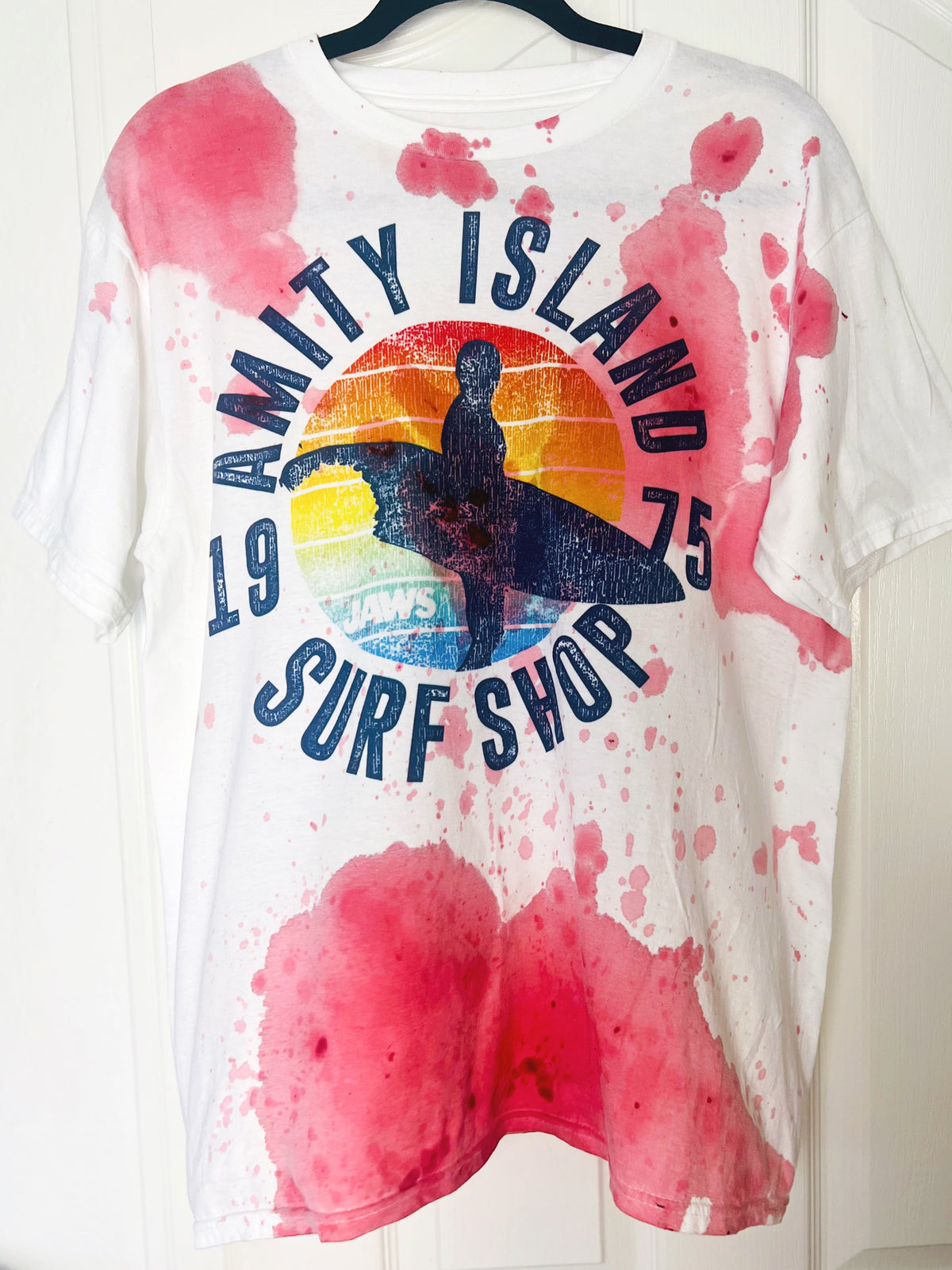 A white short-sleeve graphic T-shirt hanging on a black hanger against a white paneled door. The shirt features a distressed vintage-style print reading 'Amity Island Surf Shop' with a surfboard silhouette over a sunset circle, plus pink watercolor splatter effects around the edges.