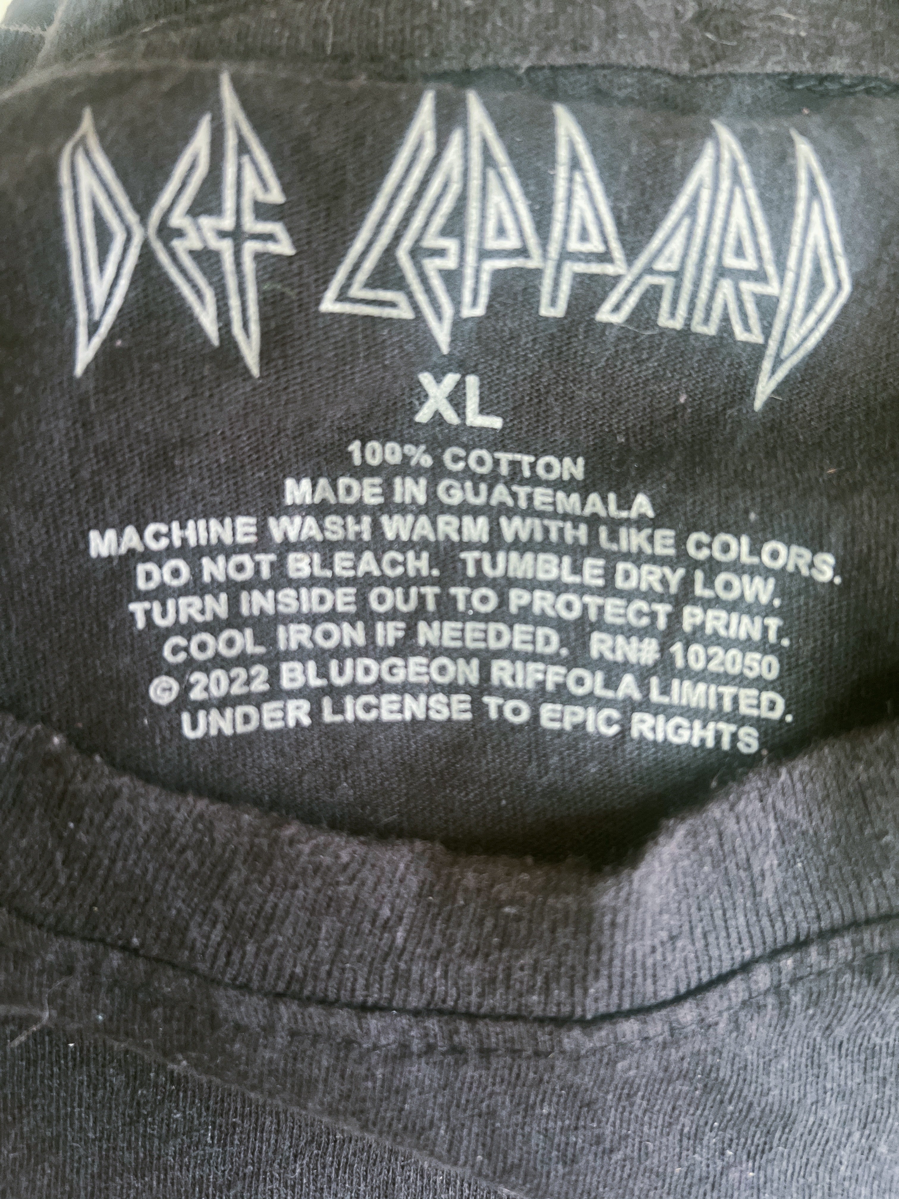 Close-up of the inside neck label of a black Def Leppard XL t-shirt, showing the white printed band logo, size 'XL', and care instructions.