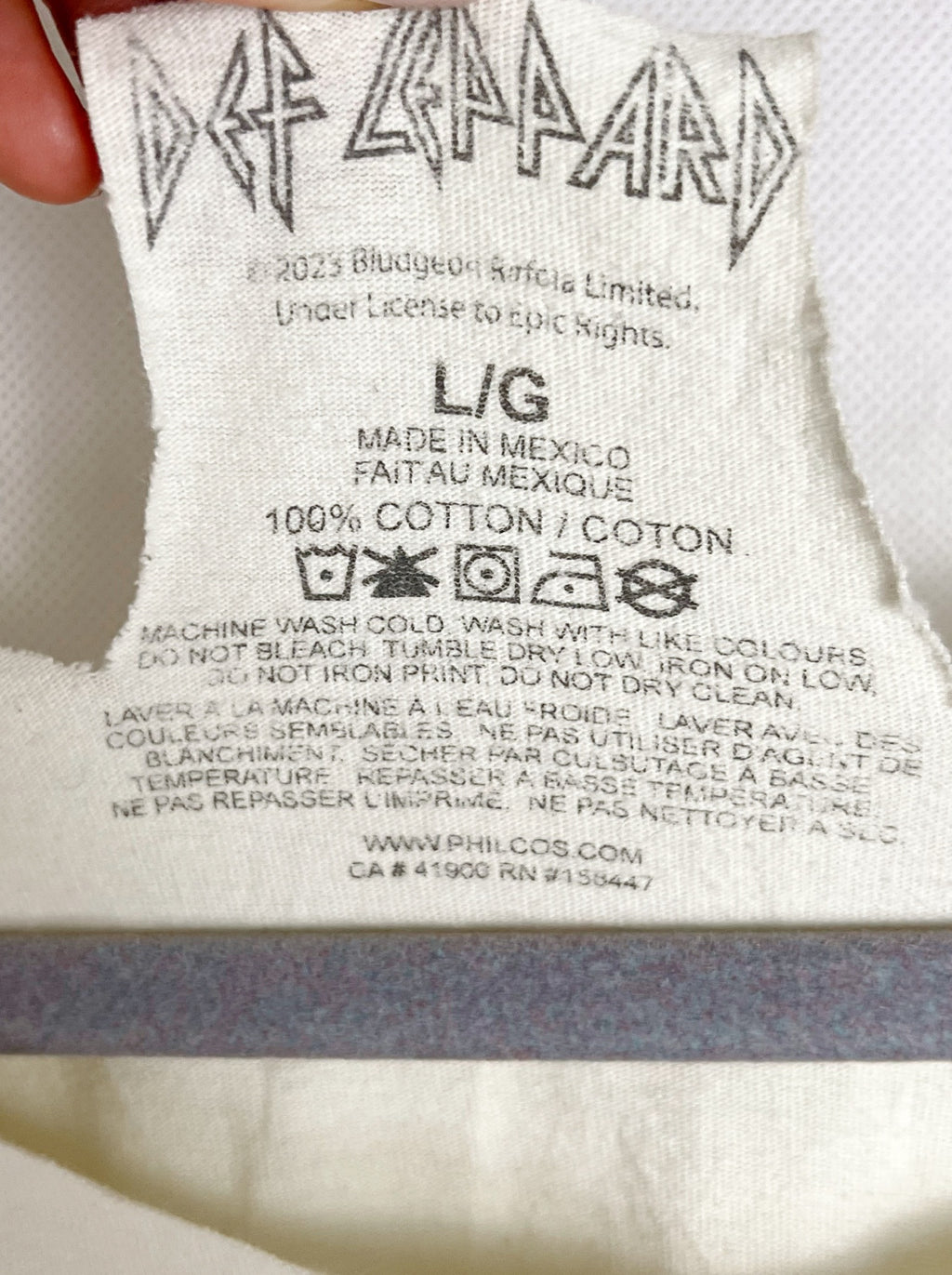 Close-up of a white fabric clothing tag with the Def Leppard band logo printed at the top, along with care instructions and size information.