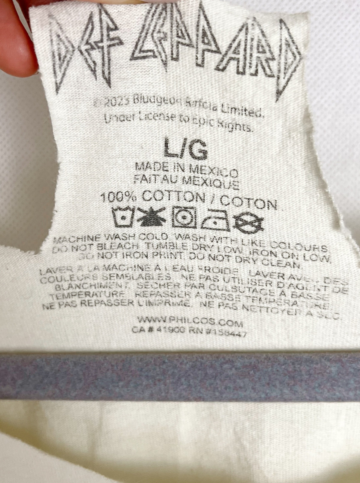 Close-up of a white fabric clothing tag with the Def Leppard band logo printed at the top, along with care instructions and size information.