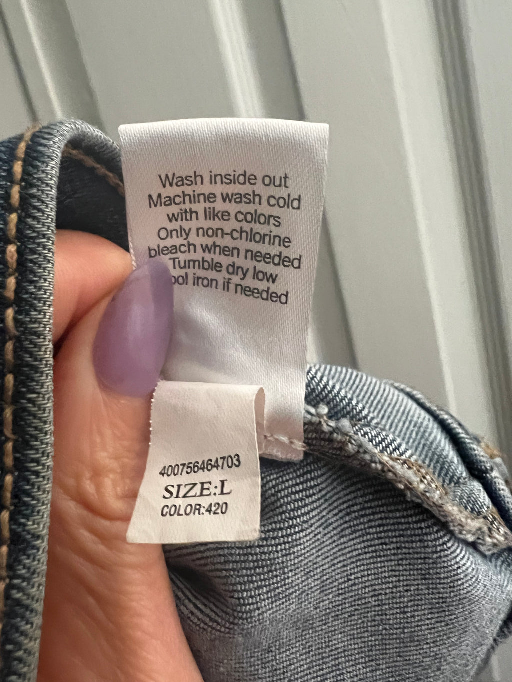A close-up of a denim garment's inner label area held by a hand with lavender nail polish, showing the care instructions and size/color tag against a light paneled background.