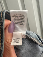 A close-up of a denim garment's inner label area held by a hand with lavender nail polish, showing the care instructions and size/color tag against a light paneled background.