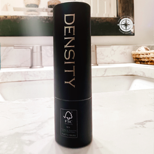 A tall black cylindrical bottle labeled 'DENSITY' with an FSC logo near the bottom, standing on a marble countertop in front of a bathroom sink and mirror.