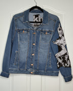 A blue denim jacket hanging on a black hanger against a white door. The jacket features a large black and white graphic print of a pin-up style woman on the right sleeve and front pocket area, with copper-toned buttons.
