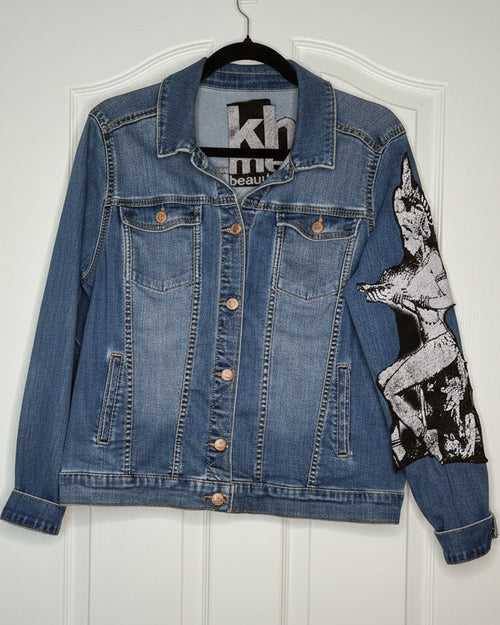 A blue denim jacket hanging on a black hanger against a white door. The jacket features a large black and white graphic print of a pin-up style woman on the right sleeve and front pocket area, with copper-toned buttons.