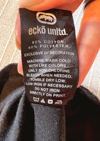 A close-up of a black fabric clothing tag held by a hand, showing the brand 'ecko unldt.' and care instructions.