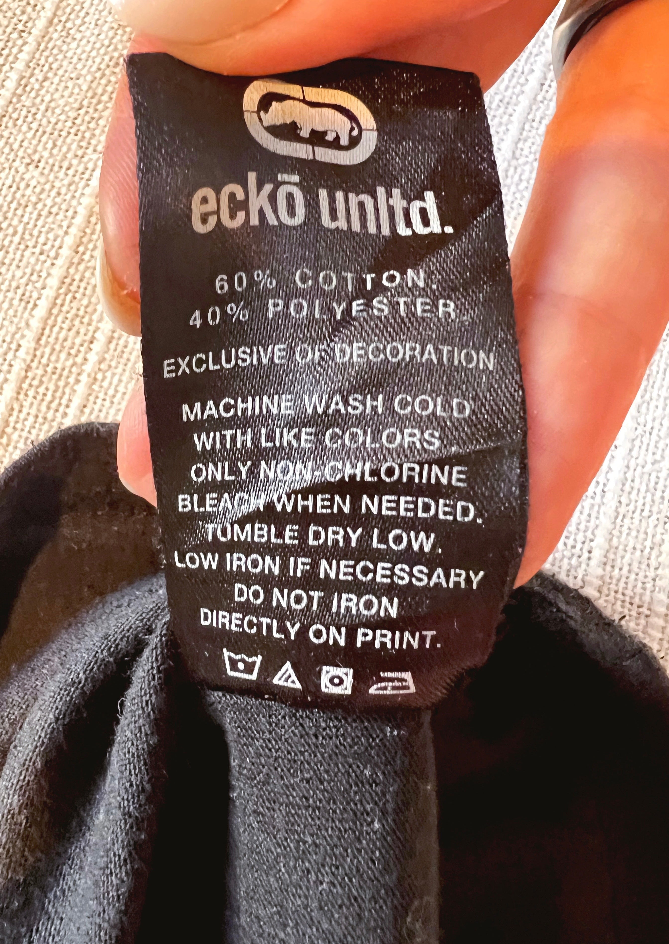 A close-up of a black fabric clothing tag held by a hand, showing the brand 'ecko unldt.' and care instructions.