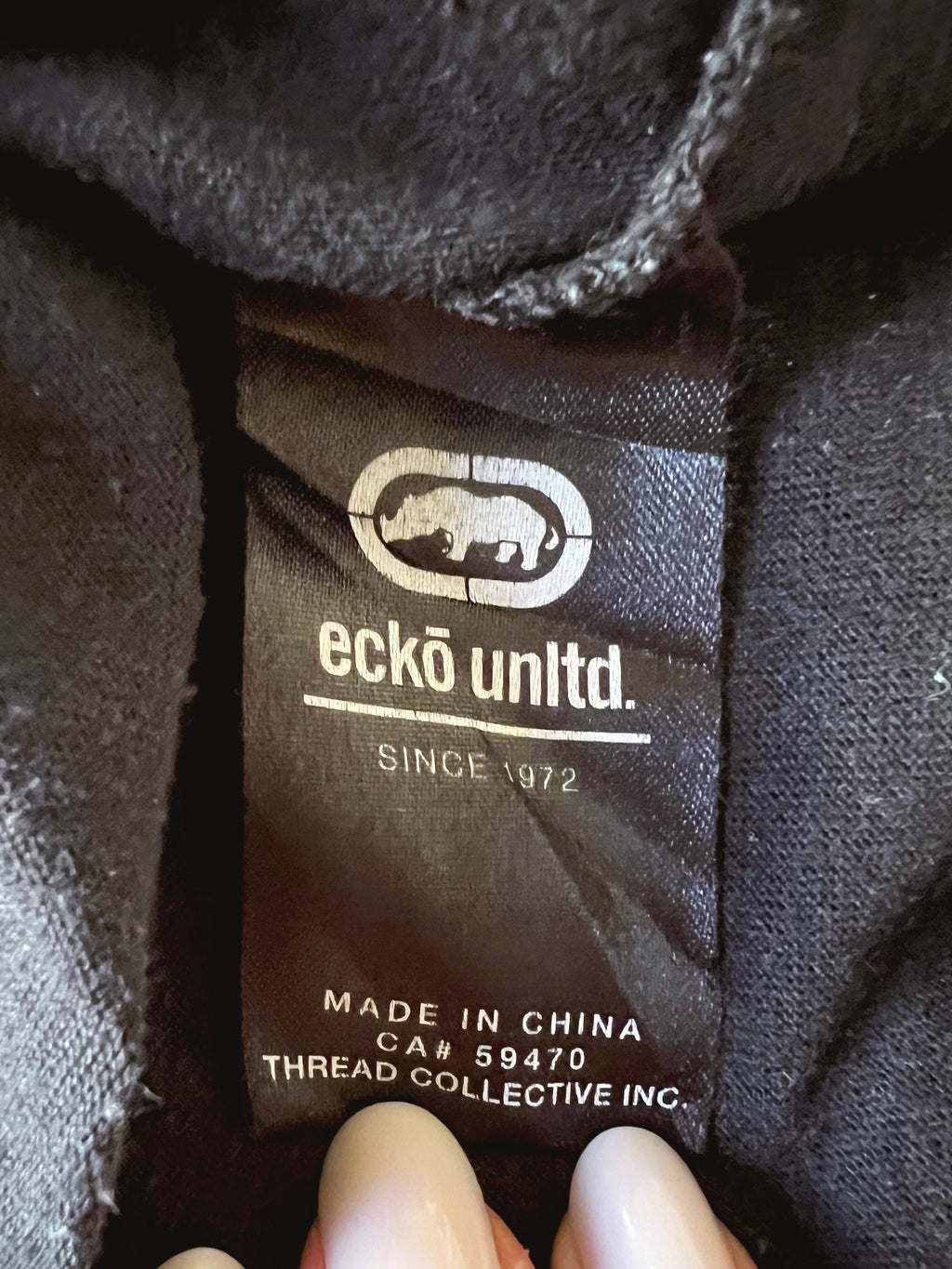 Close-up of a black clothing label reading 'eckø unldt.' with a rhino logo and 'SINCE 1972,' held by fingers against dark fabric.