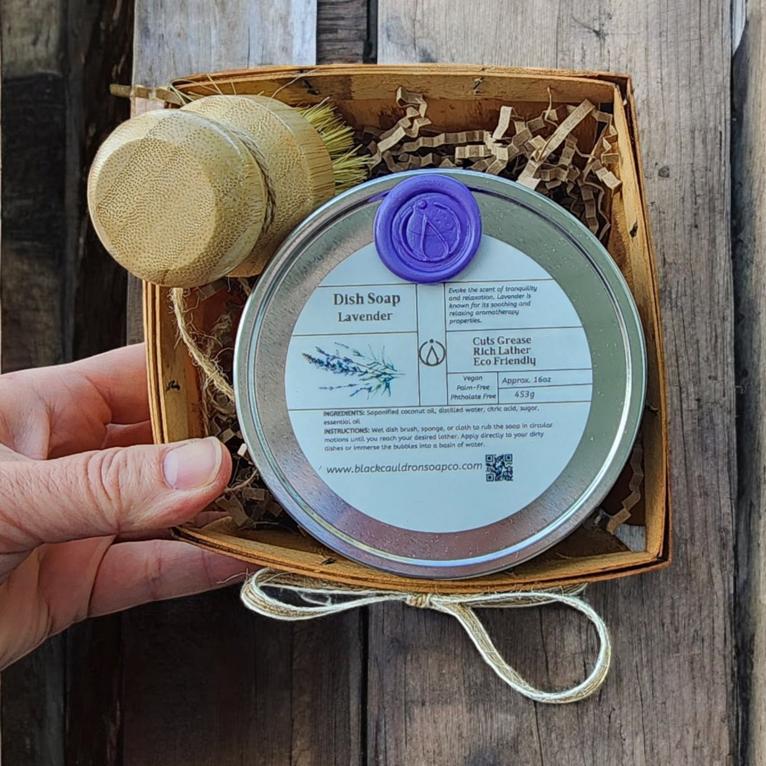 Hand holding a wooden box with a dish soap container, brush, and lavender label on a wooden surface.