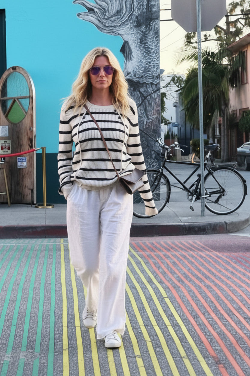 Woman in striped sweater and white pants standing on a colorful street with a blue building in the background.