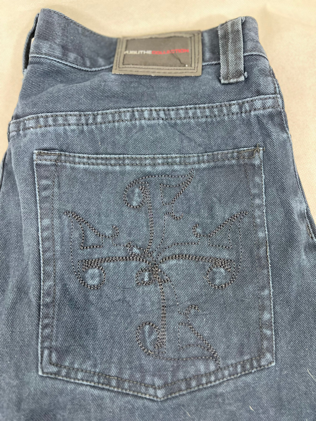 Close-up of the back of a pair of blue denim jeans, showing a large decorative embroidered cross on the back pocket and a brand label at the waistband.